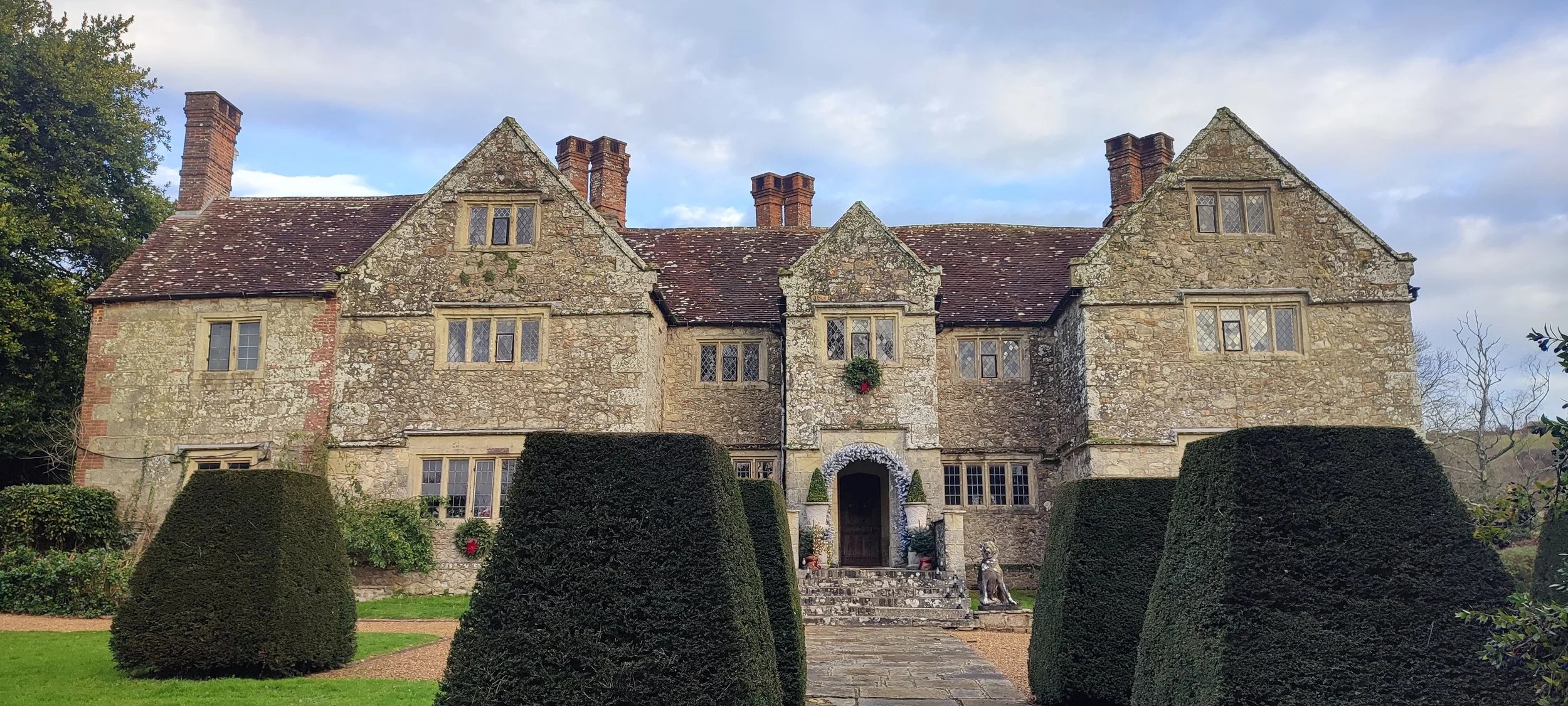 A historic stone mansion with a steeply pitched roof, multiple chimneys, and small leaded glass windows, surrounded by manicured bushes and a garden path.