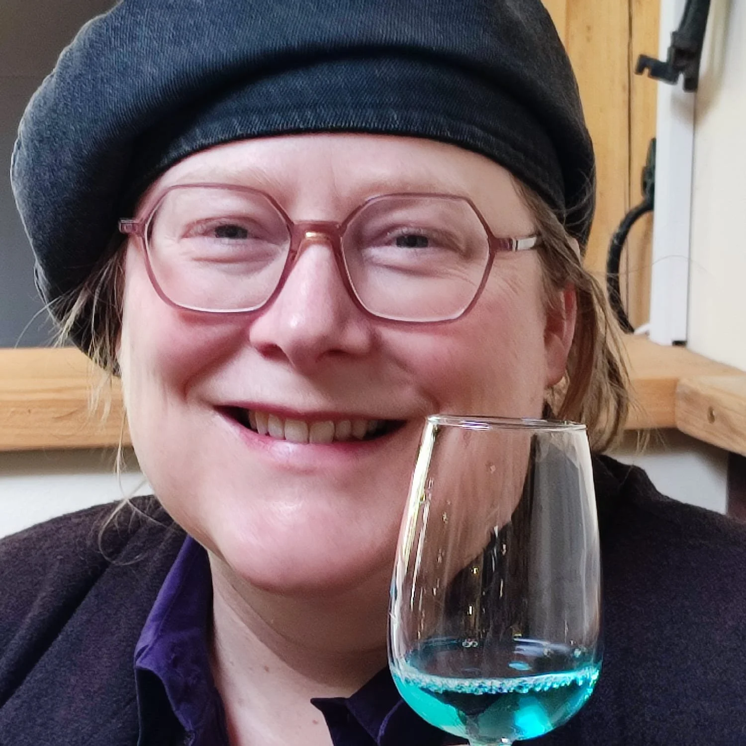 A woman with glasses and a beret holding a glass of blue-colored drink, smiling at the camera.
