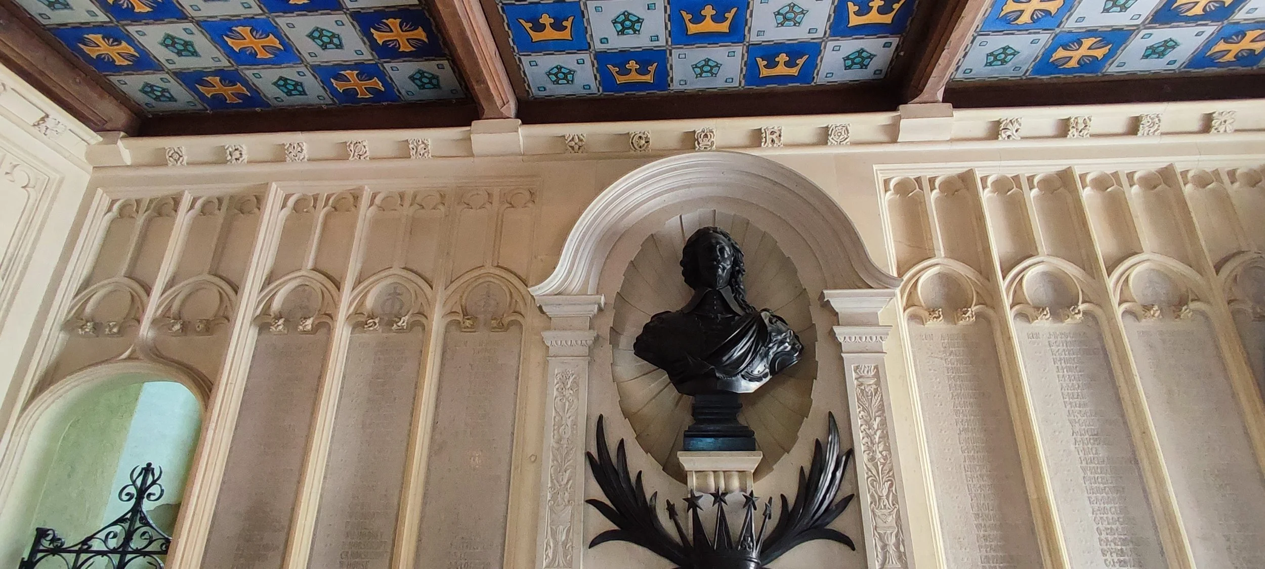 Interior of a historic building with ornate cream-colored walls, a bust sculpture of a man in the center, and a decorative ceiling with blue and gold tiles and symbols.