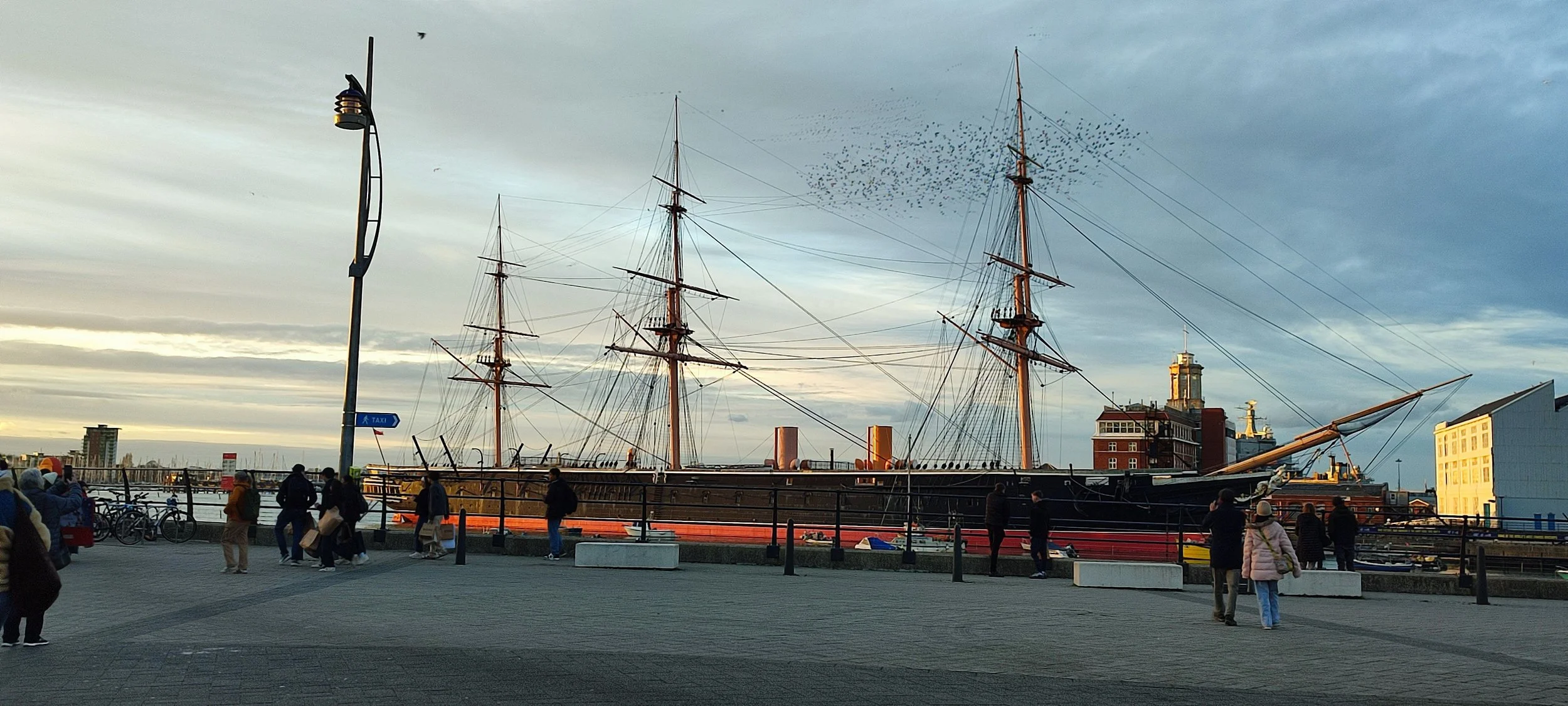 People walking along a waterfront promenade near a large historic sailing ship docked at the pier, with city buildings and cloudy sky in the background.