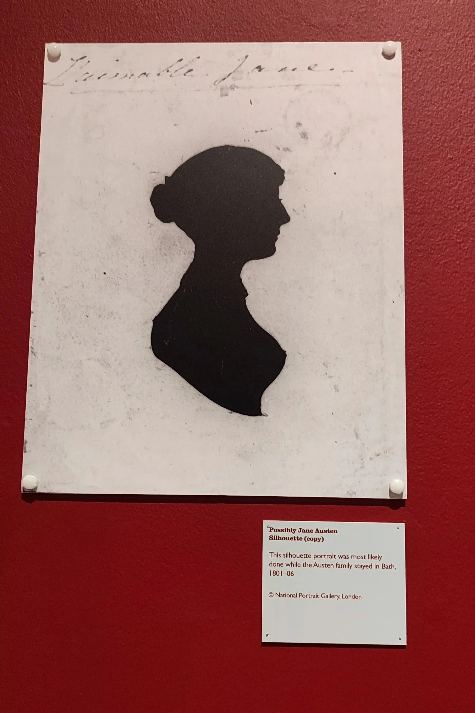Silhouette portrait of a woman with her hair in a bun, mounted on a red wall, with a caption indicating it is possibly a Jane Austen silhouette from 1801-1806.