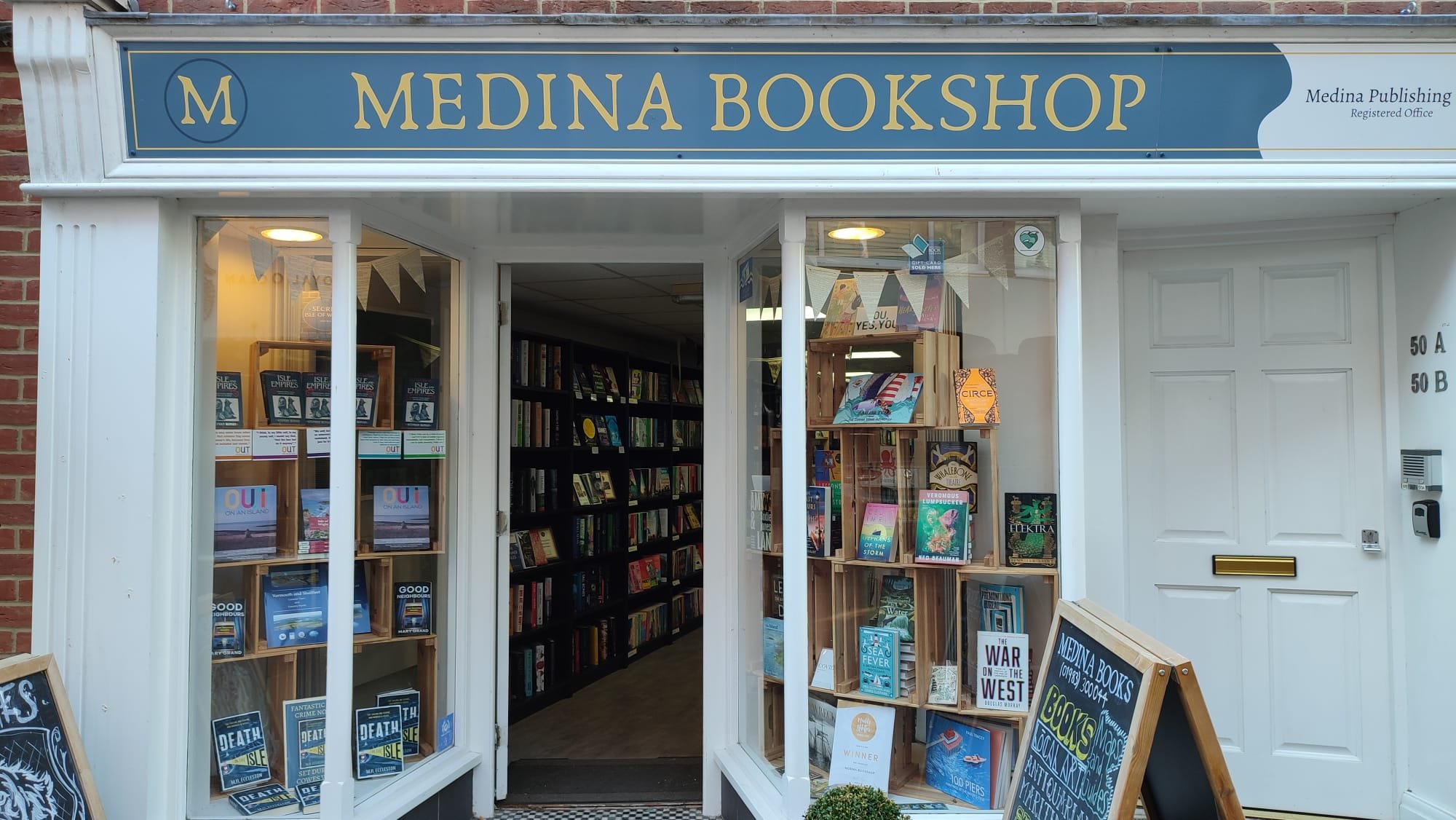 Exterior of Medina Bookshop with display windows showing books and promotional material, entrance door, and a storefront sign reading 'Medina Bookshop' in gold lettering on a blue background.