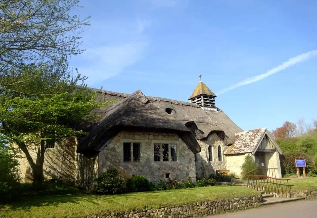 A stone church with a thatched roof, small tower, and arched windows, surrounded by greenery and a clear blue sky.
