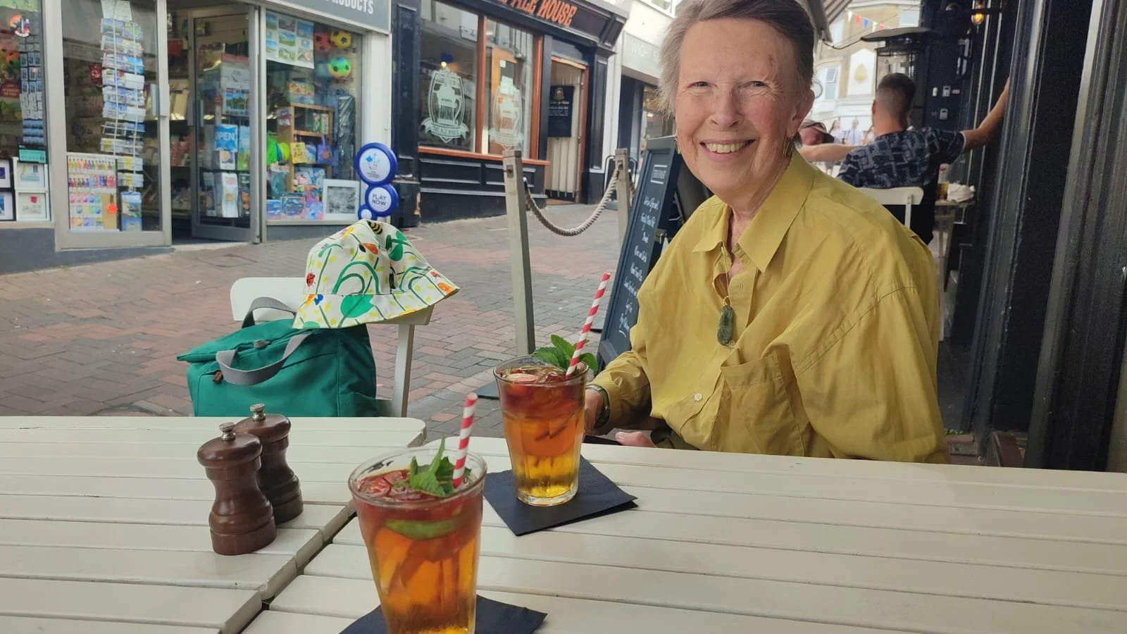 A smiling elderly woman with short gray hair wearing a yellow shirt, sitting at an outdoor cafe table with two glasses of iced tea garnished with mint and lime, and striped paper straws. The table has salt and pepper shakers, and a green backpack with a colorful hat hanging on a nearby chair. In the background, there are storefronts and a few people walking.