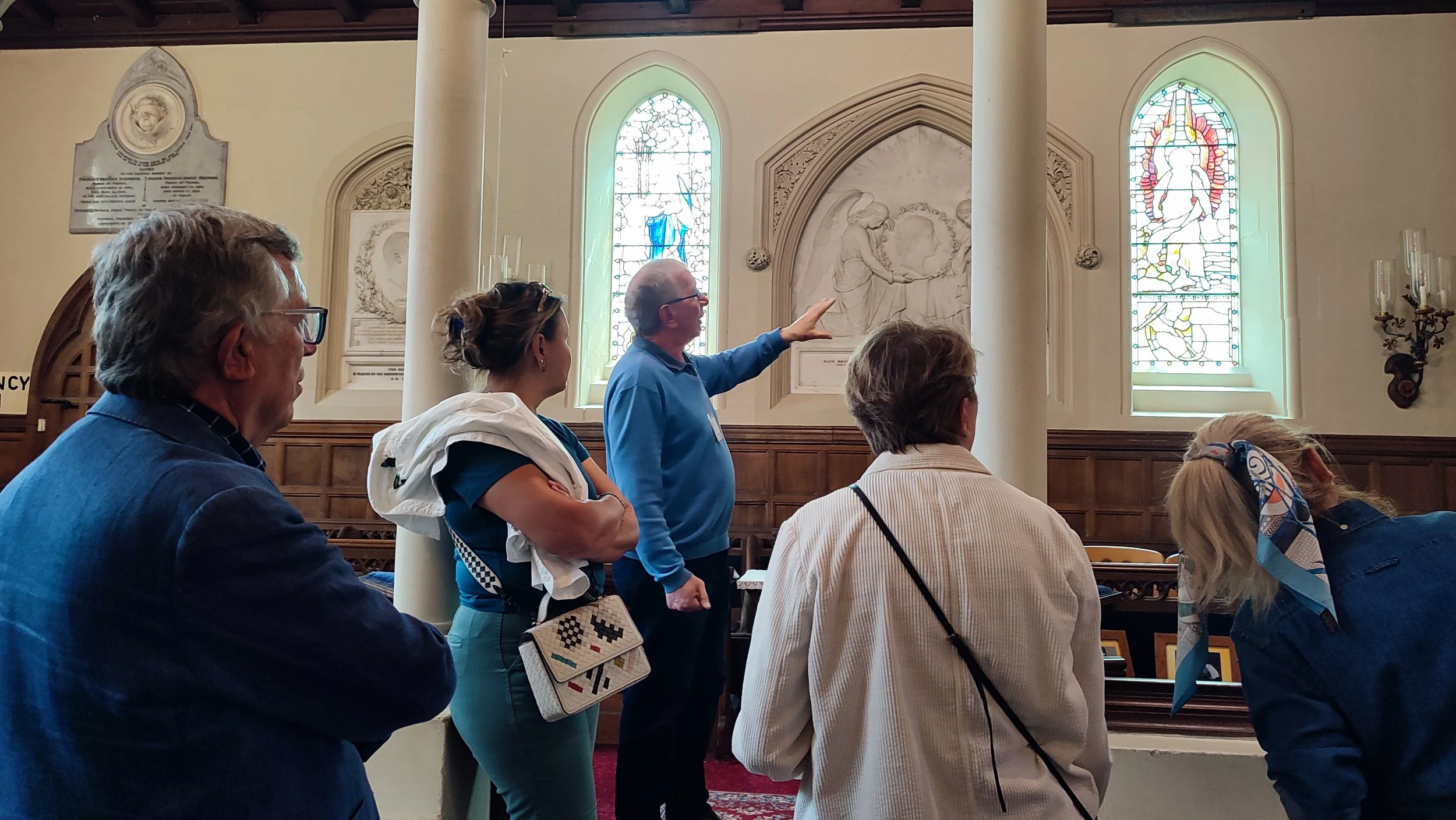 A group of five people, three women and two men, are inside a church looking at a guide who is pointing to a wall with stained glass windows and religious artwork behind them. The guide is an older man wearing glasses and a blue shirt. The church interior features stained glass windows, wooden paneling, and wall decorations.