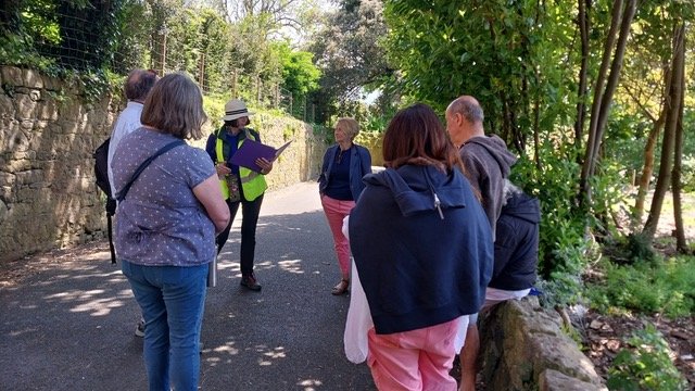 Group of people in outdoor setting, standing on a paved path, listening to a woman with a hat and high-visibility vest who is holding a notebook, surrounded by green trees.