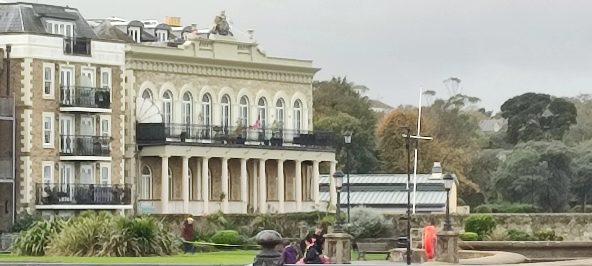 A multi-story historic building with balconies and arched windows, with people sitting and walking in front of it, in a park-like setting with trees and street lamps.
