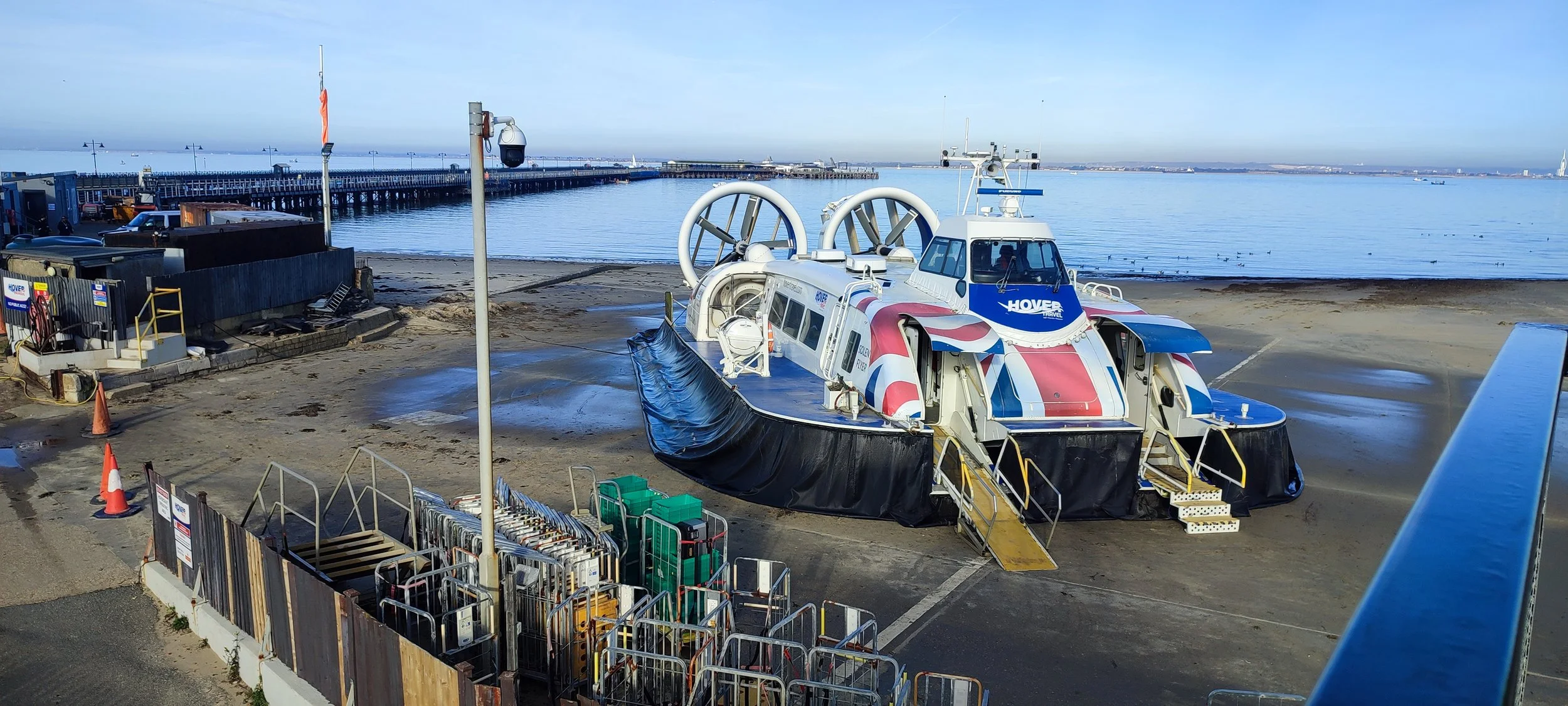 Hovercraft on a sandy beach with a pier and ocean in the background, equipment storage, and traffic cones.