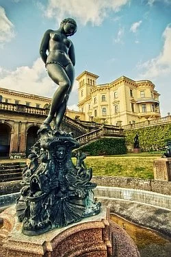 A bronze statue of a young boy standing on a fountain with a large, ornate building in the background.
