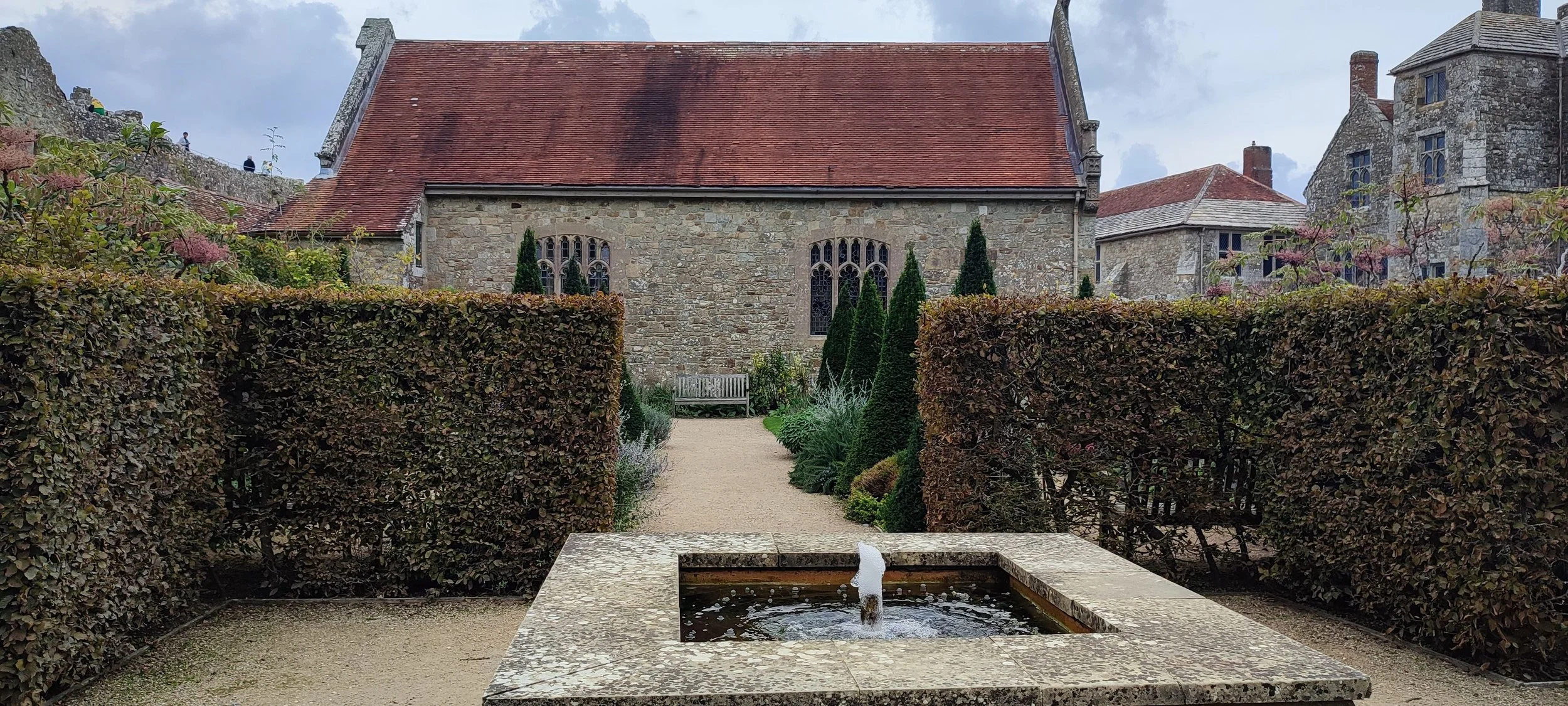 A stone pathway leading to a church with a red roof, surrounded by manicured hedges and tall, narrow trees, with a fountain in the foreground.