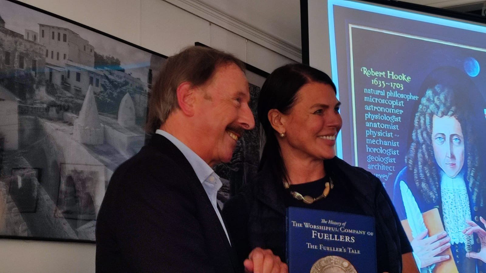 Two smiling individuals, a man with gray hair and a woman with dark hair, are standing together at an event. The woman holds a blue book with gold embossing. Behind them, a large screen displays a slide about Robert Hooke, listing his professions and accomplishments. A framed black-and-white photograph of a cityscape is on the wall behind them.