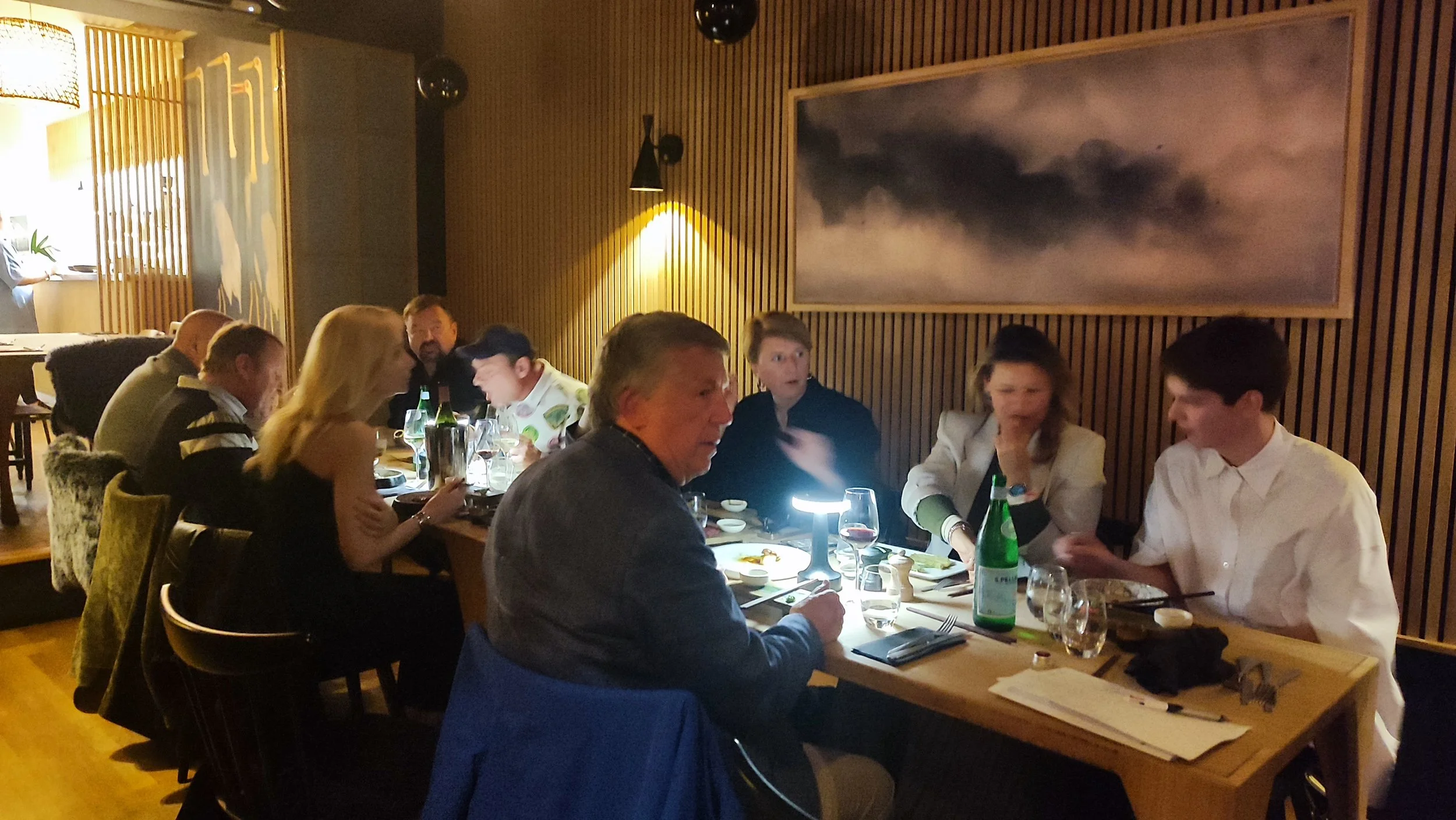 People dining at a restaurant table in a cozy, dimly lit atmosphere with wooden wall decor and a large abstract painting.