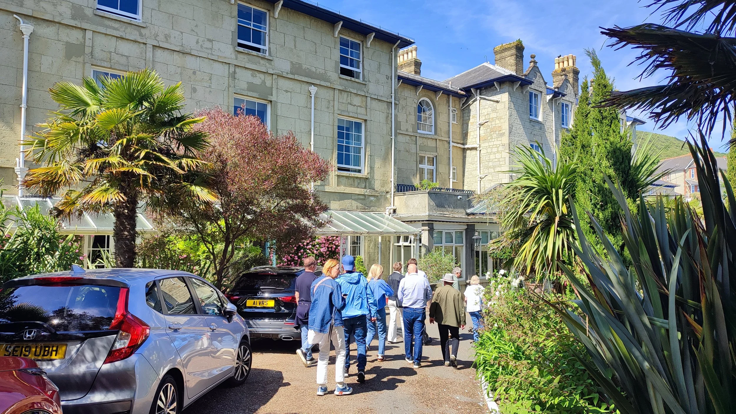 A group of people on a tour walking on a gravel pathway in front of a large, historic stone building with multiple windows and a glass conservatory attached at the entrance, surrounded by lush greenery and tall trees.