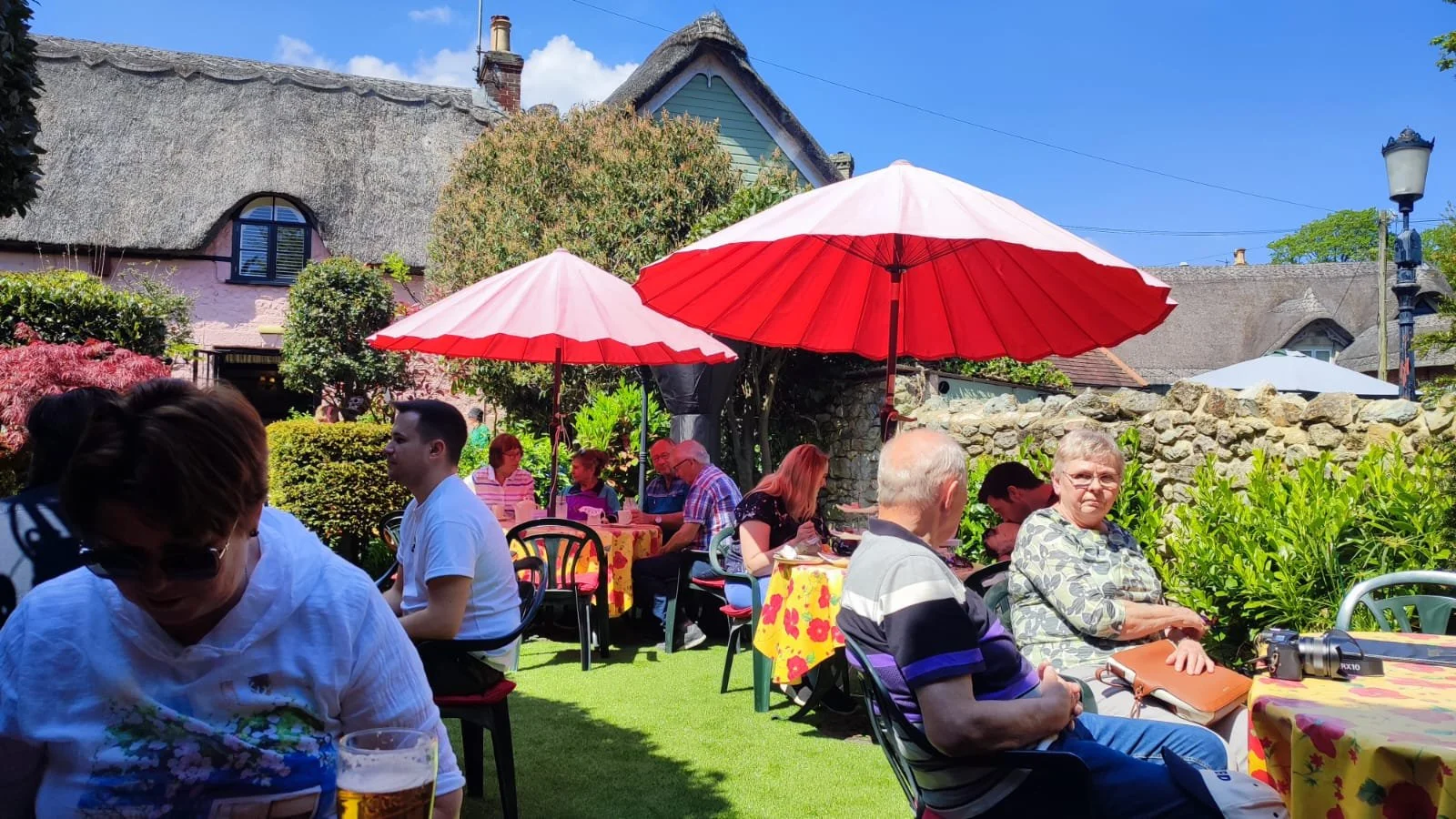 People sitting at outdoor tables under red and white umbrellas in a garden on a sunny day, with traditional buildings and greenery in the background.