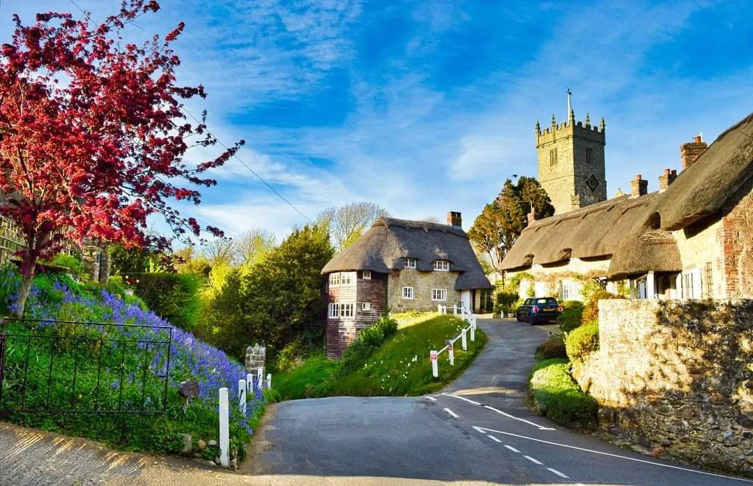 A picturesque village scene with a narrow road leading to historic thatched-roof cottages and a stone church tower. Brightly colored flowers and lush green trees surround the buildings on a sunny day.
