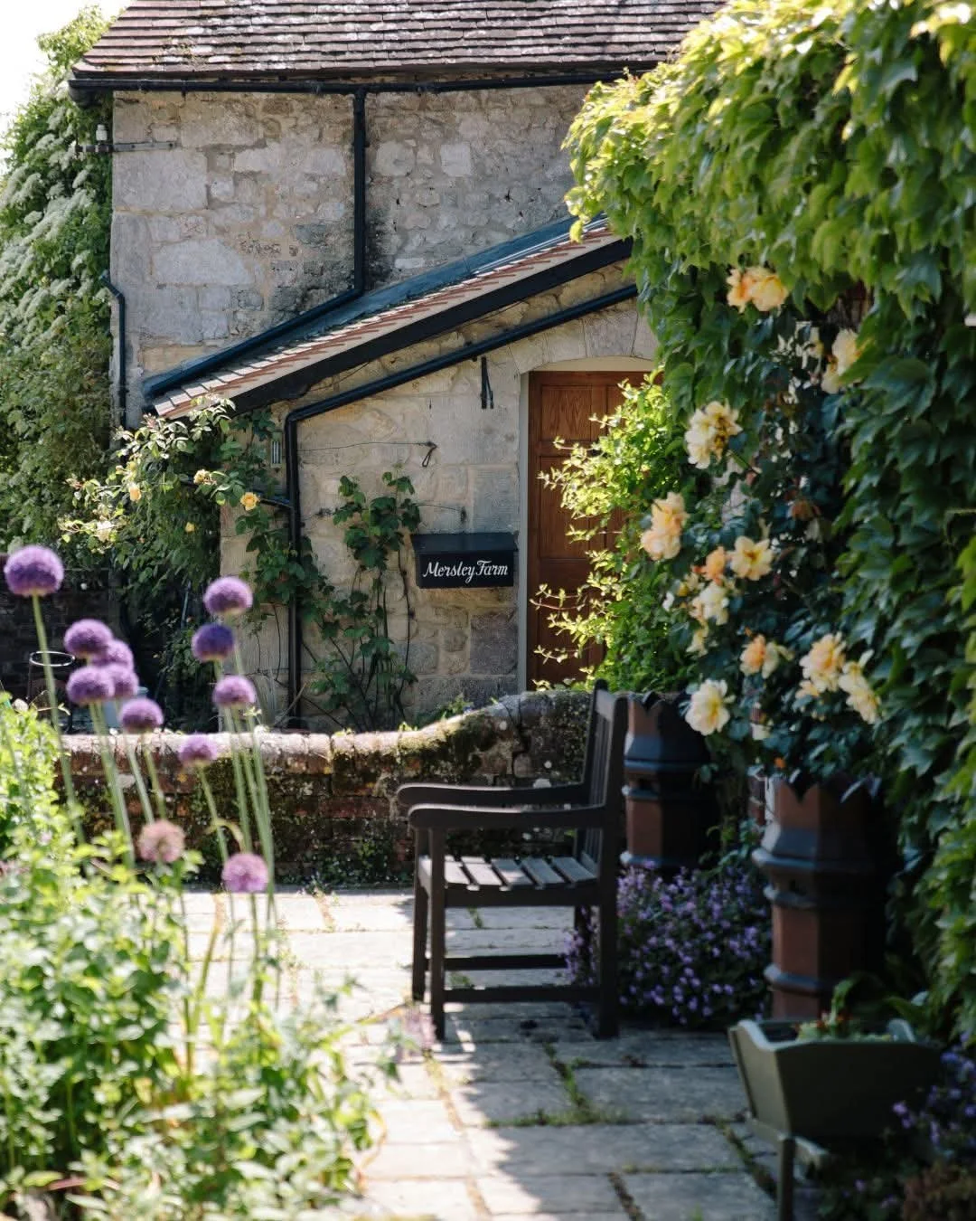 A cozy garden patio with a stone building in the background. The patio has a black bench, potted plants, and blooming flowers, including purple alliums and yellow hibiscus, surrounded by lush greenery and climbing vines. There is a sign on the wall that says 'Mersley Farm'.