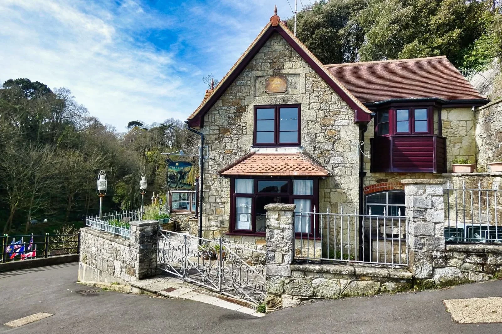 A stone house with red-tiled roof and purple window frames, surrounded by a white metal fence, with a hilly background and trees.