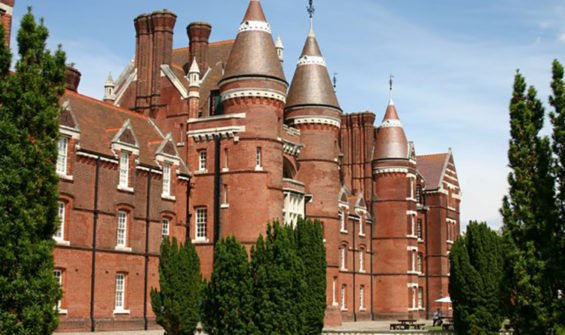 A large red brick castle with multiple towers and turrets, surrounded by green trees under a clear blue sky.
