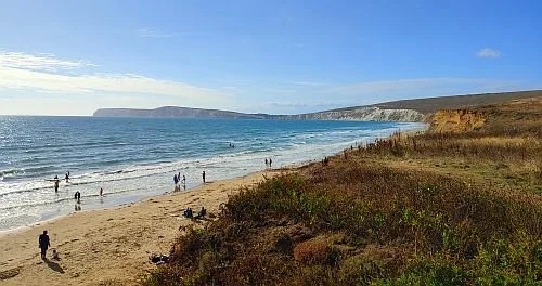 Beach scene with people walking and relaxing, ocean waves, cliffs in the background, clear sky.