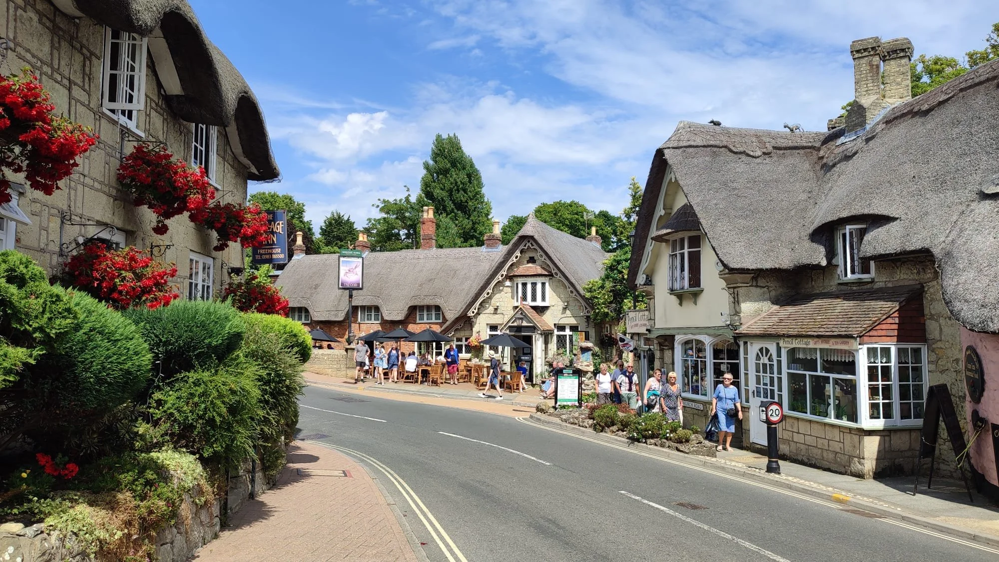 A quaint street scene in a village with thatched-roof buildings, blooming red flowers, and tourists walking and sitting outside cafes on a sunny day.