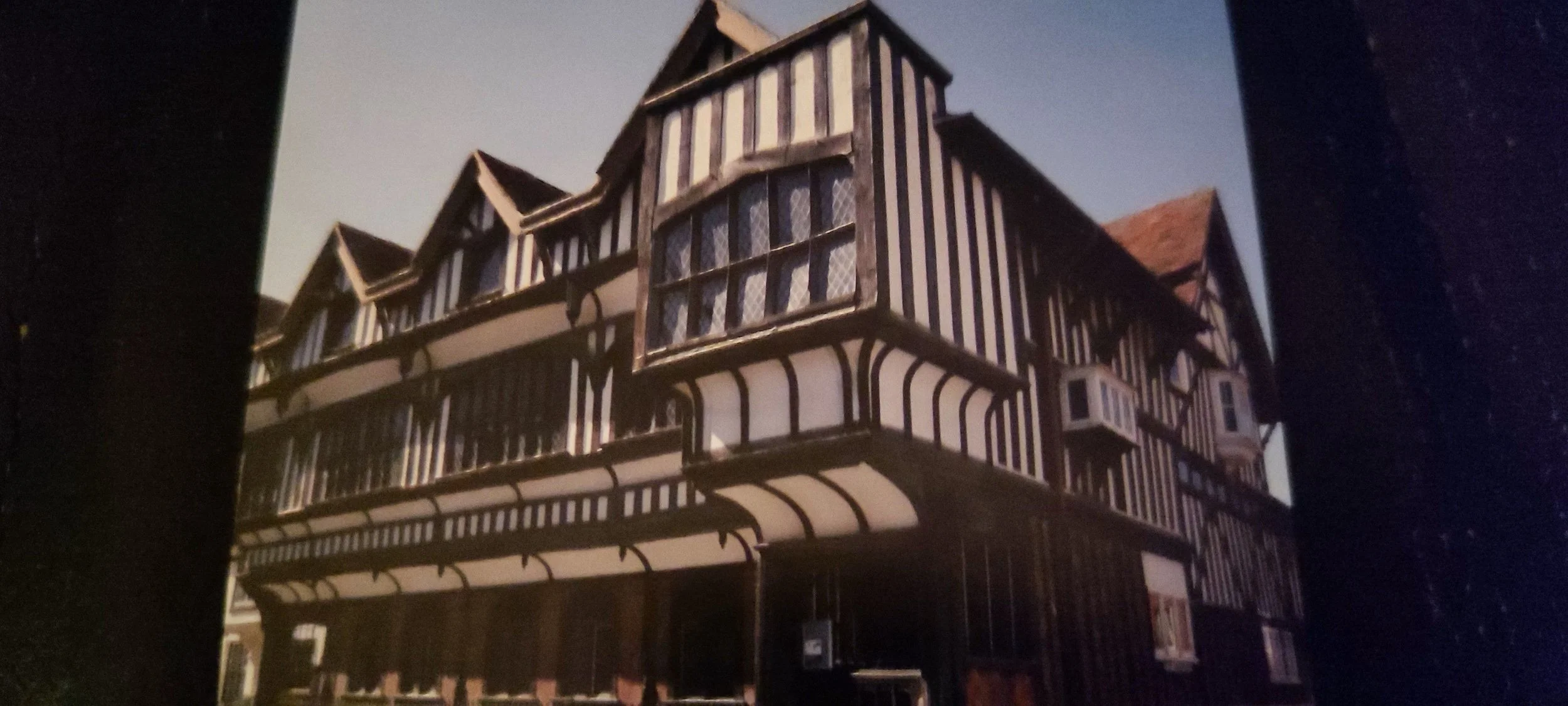 A multi-story building with a Tudor-style facade, featuring black and white vertical and horizontal wood beams, multiple windows, a large bay window on the front, a gated entrance, and part of a red-tiled roof visible at the top.
