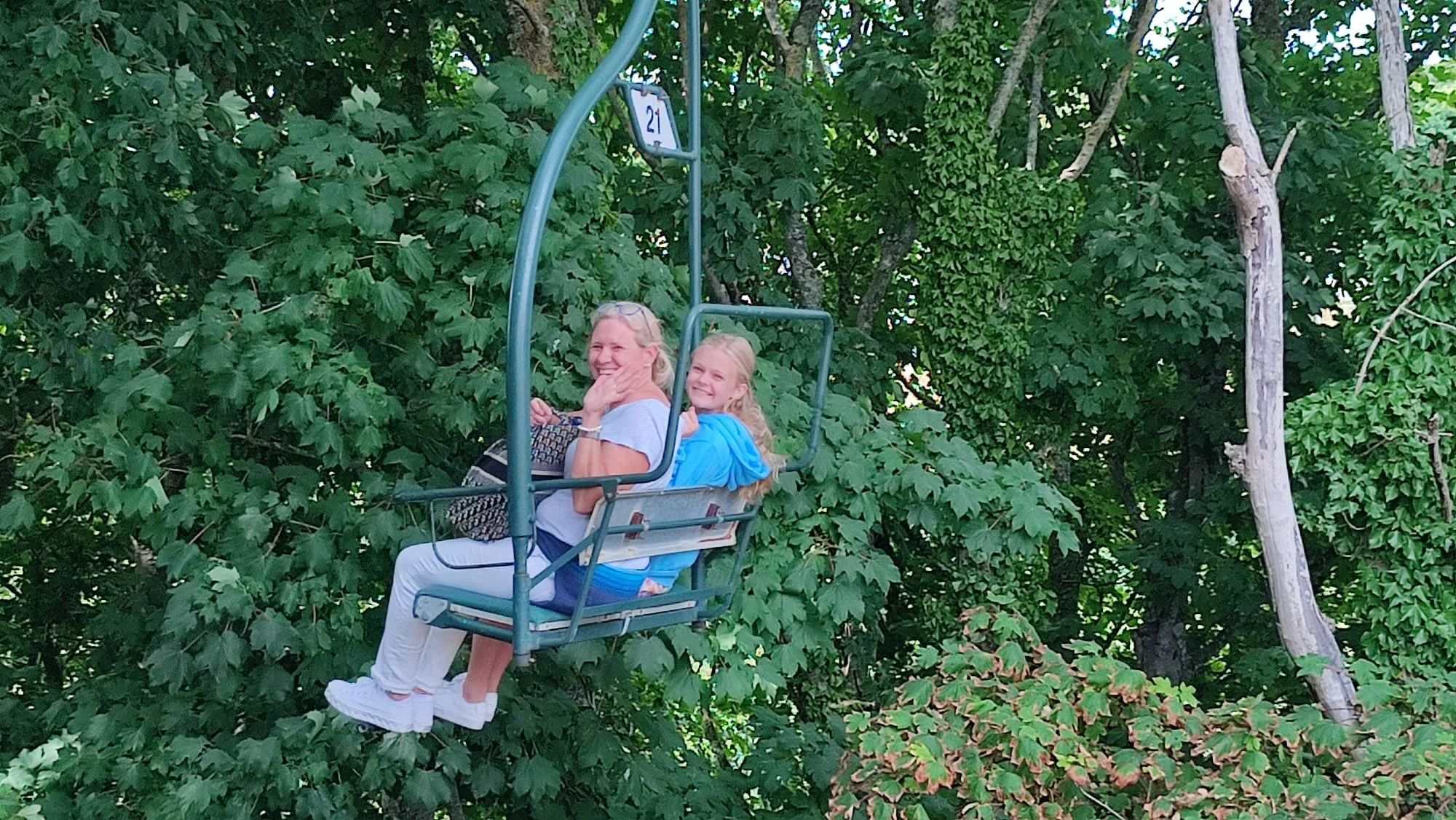 Two light-skinned, smiling women and a girl sitting on a ski lift, surrounded by green leafy trees.