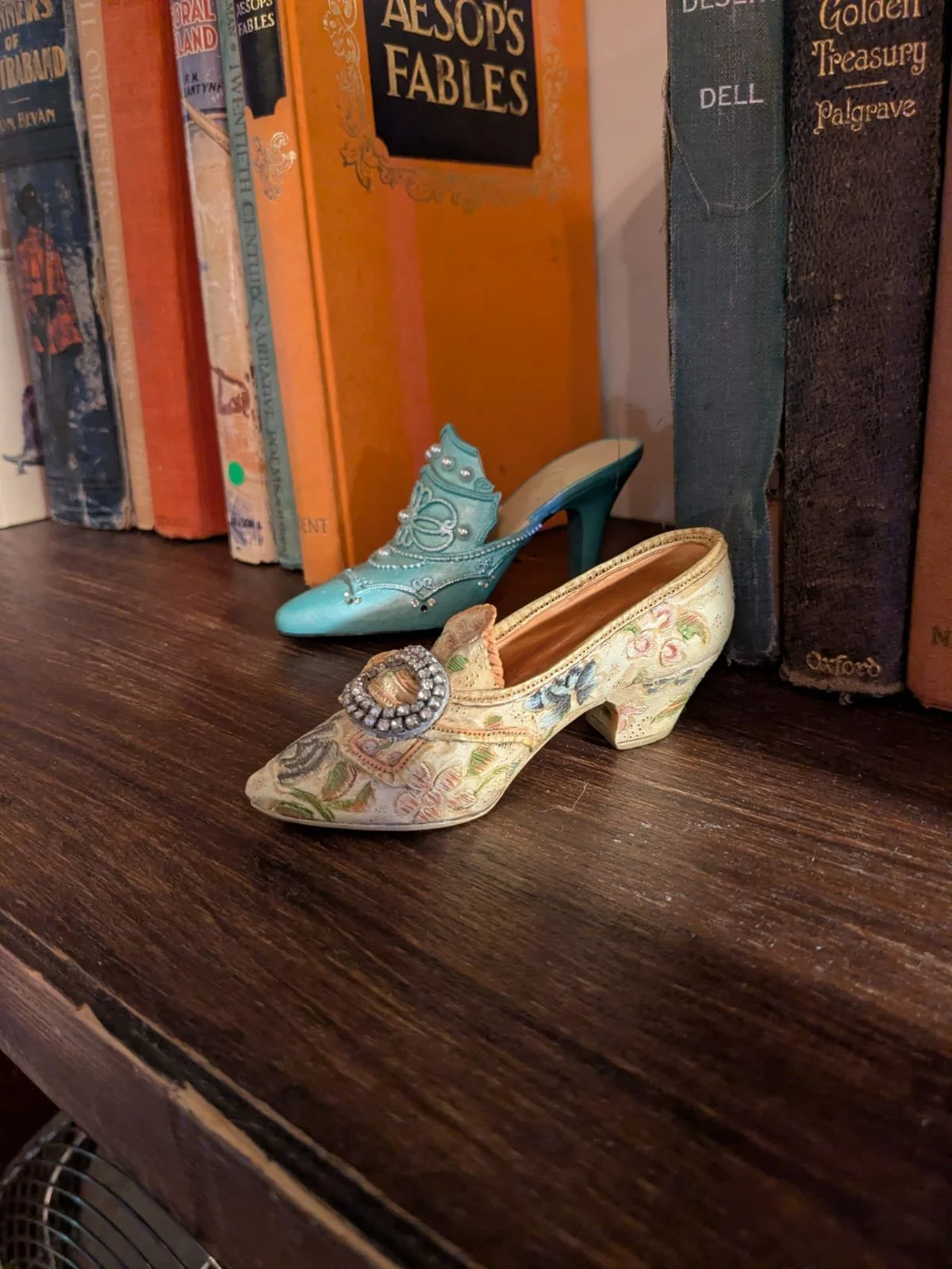 Two decorative vintage-style shoes on a wooden shelf in front of a row of colorful books.