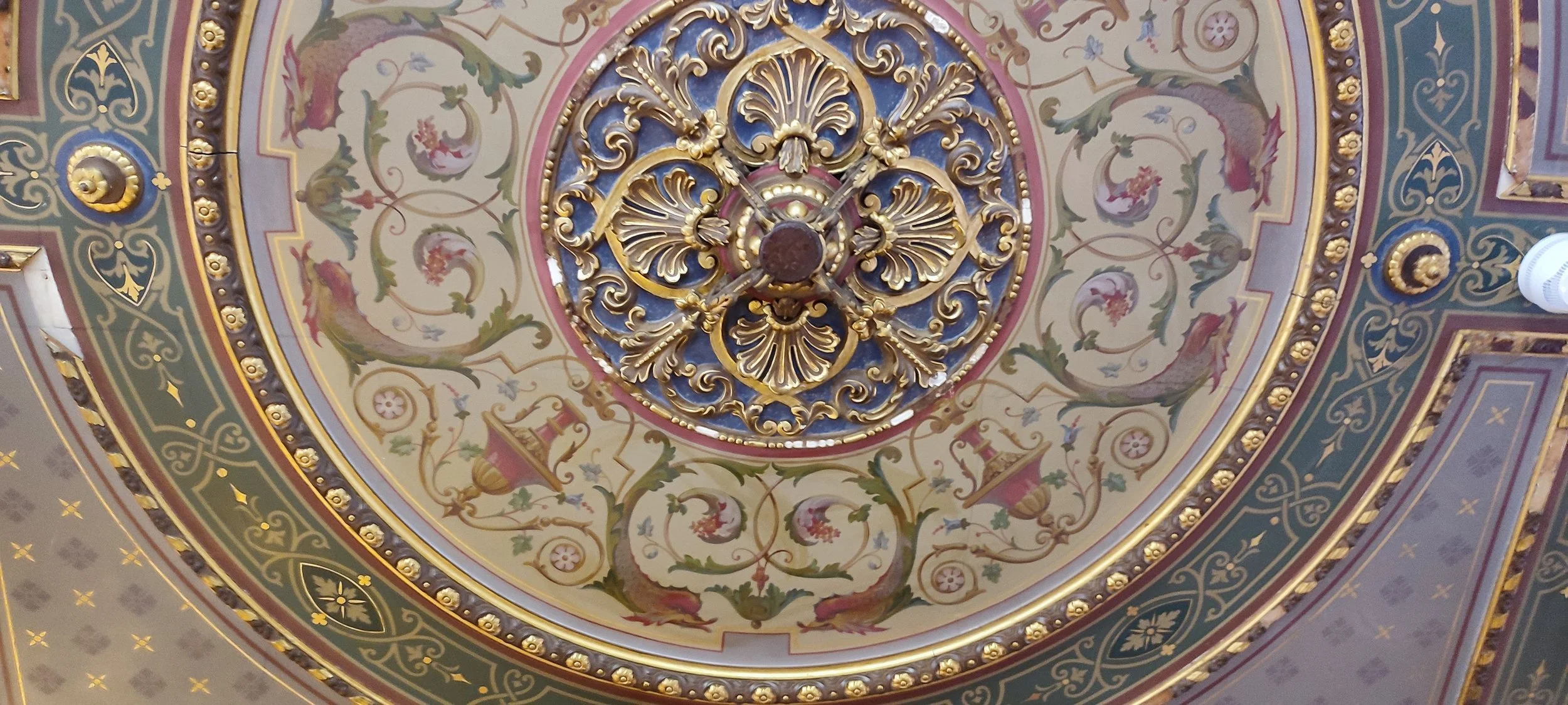 Ornate ceiling with a gold decorative chandelier at the center, painted with floral designs, scrolls, urns, and mythological creatures, featuring blue, green, red, and gold accents.