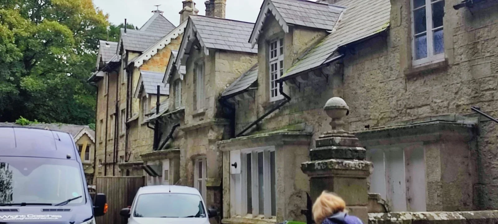 Stone row of old houses with steep gabled roofs, some with dormer windows and black drains, and trees in the background. Two cars are parked in front, and a person with blonde hair is looking at the buildings.