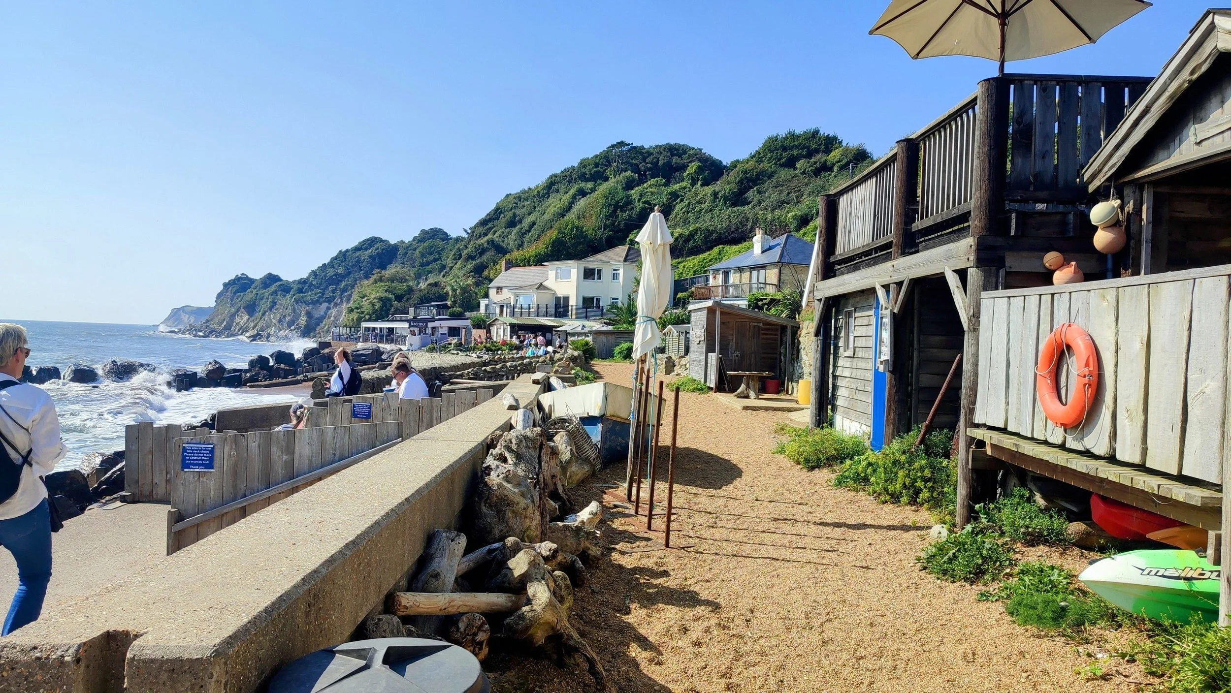 Beachside walkway with wooden fencing and buildings, people sitting near the ocean, with a lush green hillside in the background under a clear blue sky.