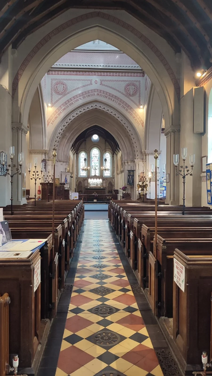 Interior of a church with wooden pews, stained glass windows, and an altar at the front. Ornate chandeliers and decorative elements are visible, with a patterned tile floor leading up to the altar.
