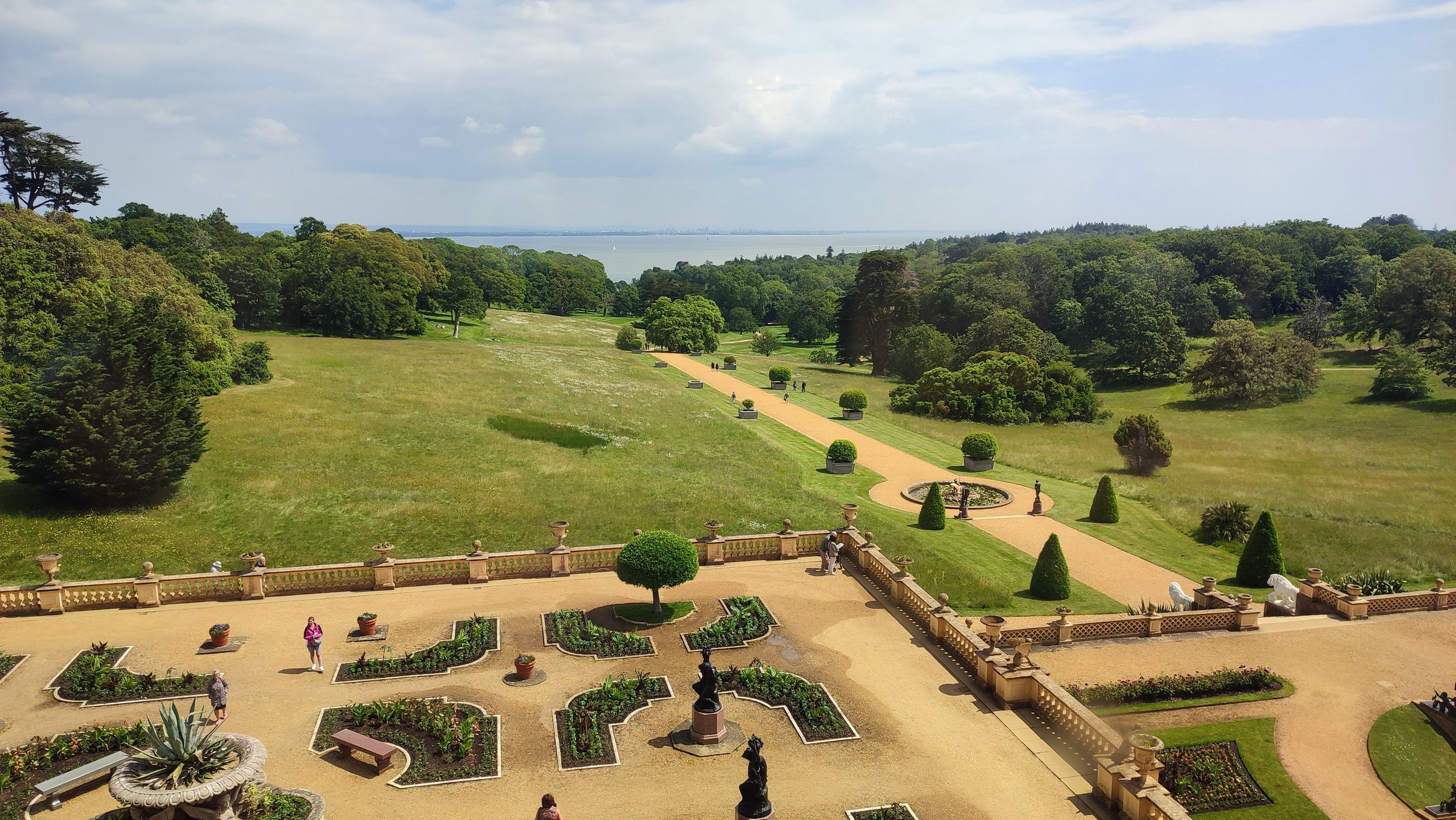 A grand outdoor garden with pathways, sculptures, and well-maintained greenery, overlooking a scenic body of water and lush trees in the distance.