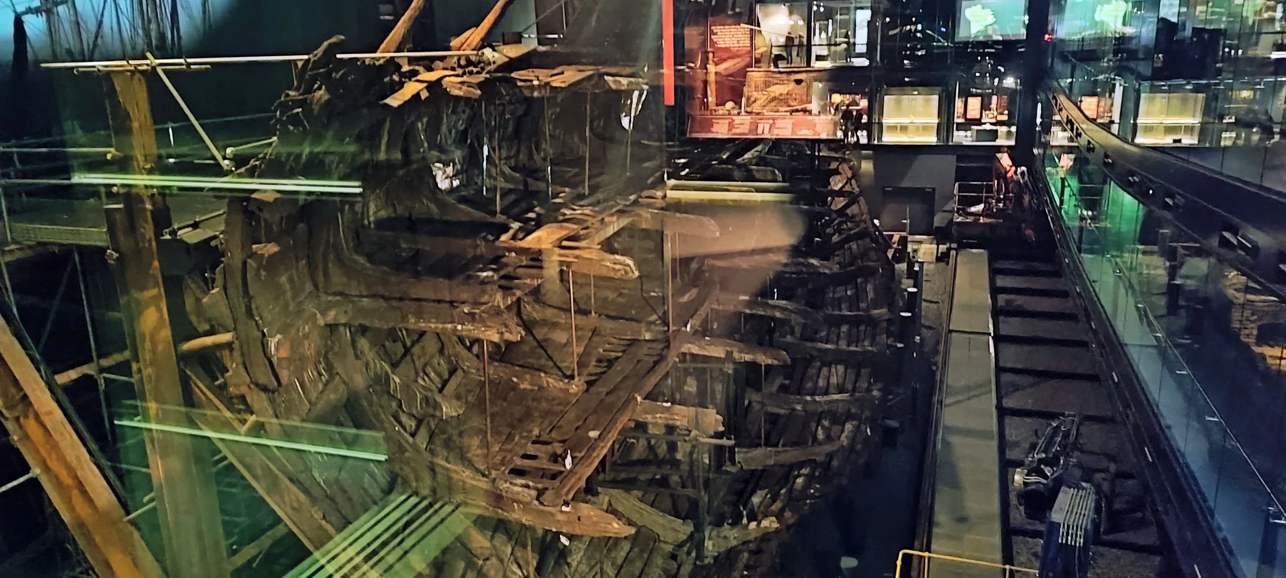 View of a boat wreck inside a glass display at a museum, with modern walkways and exhibits surrounding it.