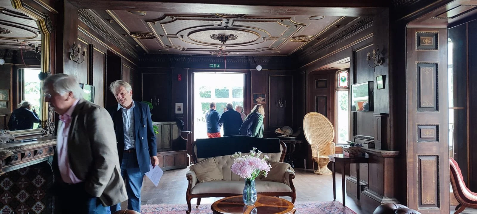 Interior of a room with dark wooden walls, ornate ceiling, a couch with a vase of pink flowers, a mirror, a television, and several people standing and talking near the entrance.