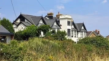 A hillside with lush green grass and shrubs, topped by several large, historic houses with dark roofs and ornate architecture under a blue sky.