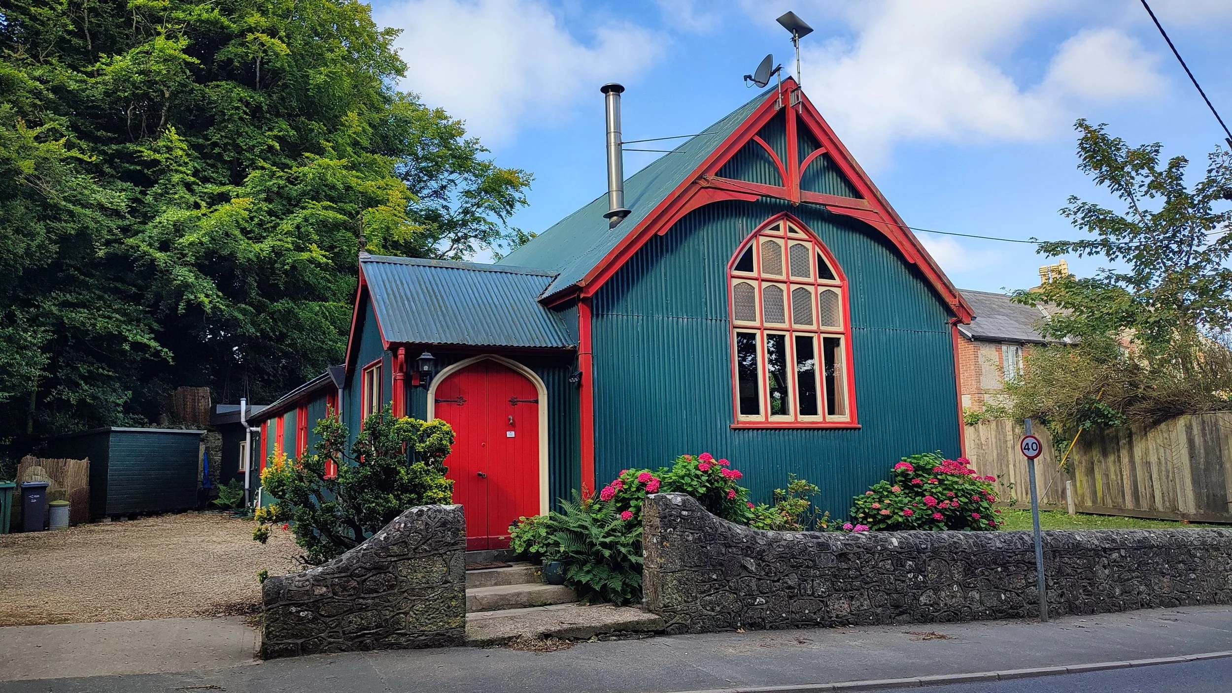 A colorful church with green corrugated metal walls, red trim, and a red door. It has large arched windows, a stone staircase entrance, and is surrounded by flowering bushes. The sky is partly cloudy, and there are trees and a fence nearby.
