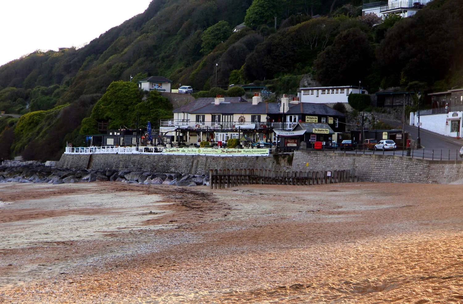 Beachfront restaurant and inn with outdoor seating, positioned along a rocky waterfront with a retaining wall, with houses and hillside covered in trees in the background.