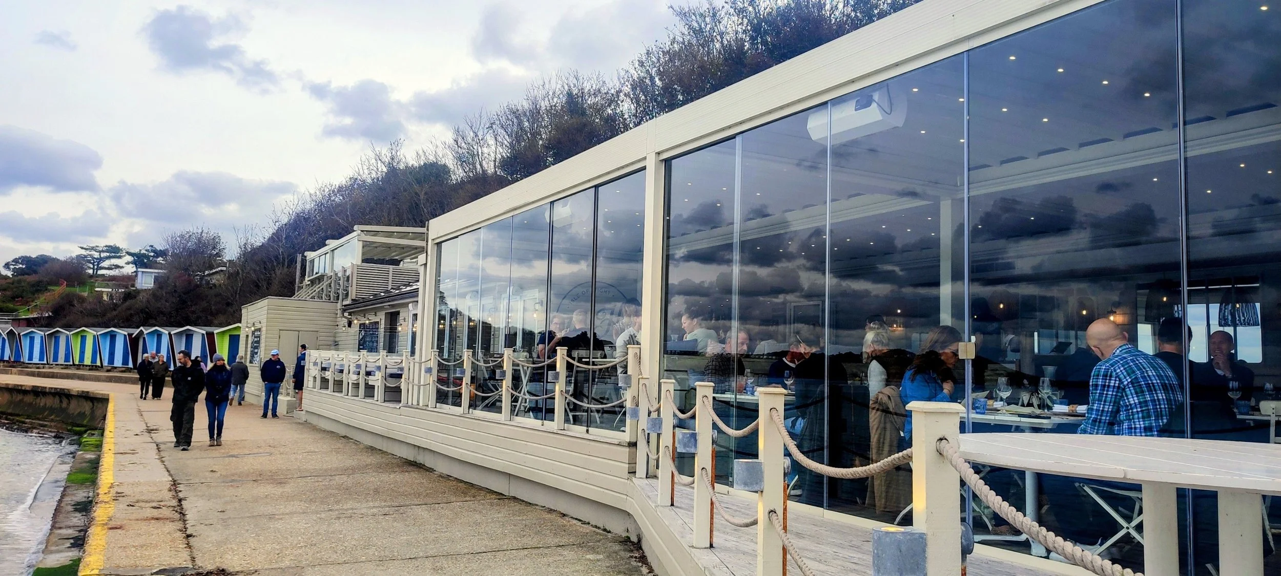 People dining inside a modern restaurant with large glass windows, overlooking a cloudy sky and surrounding landscape, with some customers standing outside near the entrance.