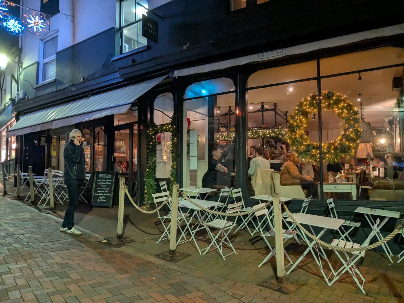 Exterior view of a restaurant decorated for Christmas with wreaths and string lights. A person is standing outside on the sidewalk talking on a cellphone, and there are empty outdoor tables and chairs in the foreground. Inside, people are dining behind large glass windows.