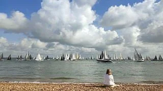 A person sitting on a beach facing a large body of water with numerous sailboats in the distance under a cloudy sky.
