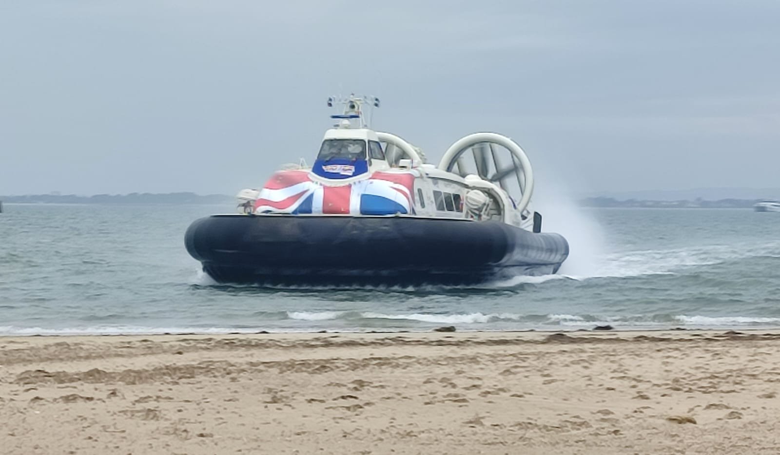 Hovercraft with a Union Jack flag design on the front, moving over water near a sandy shore.