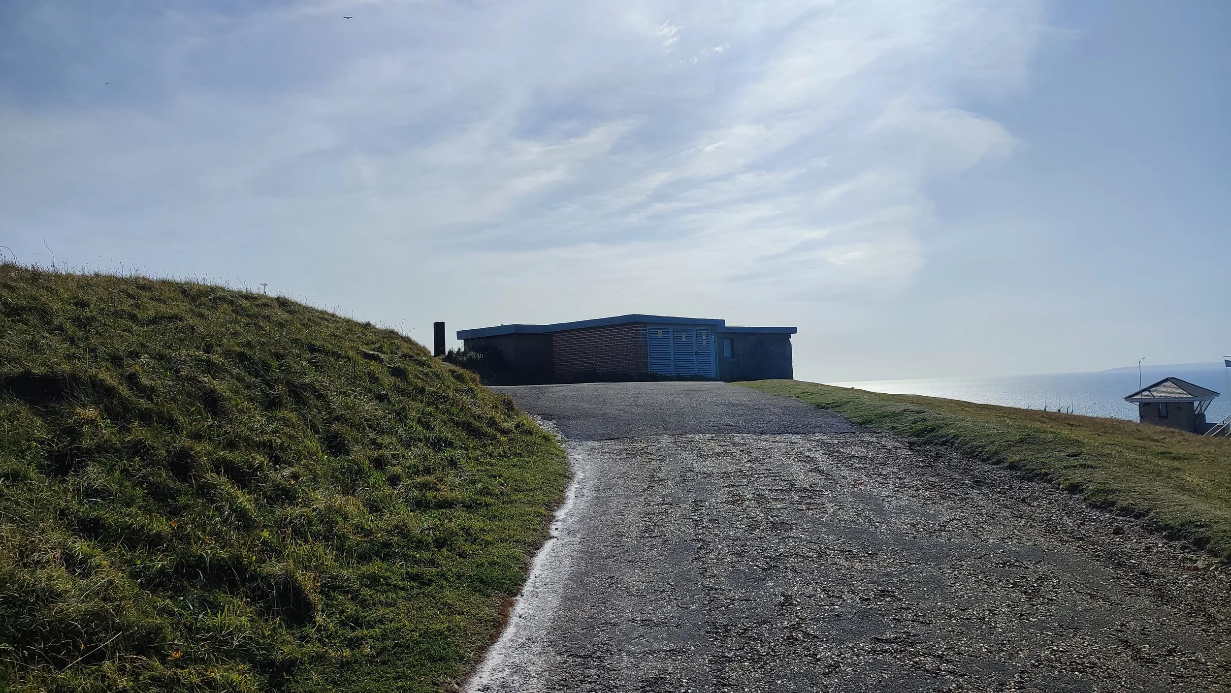 A gravel path on a grassy hill leading to a small building with blue doors, overlooking the ocean on a partly cloudy day.