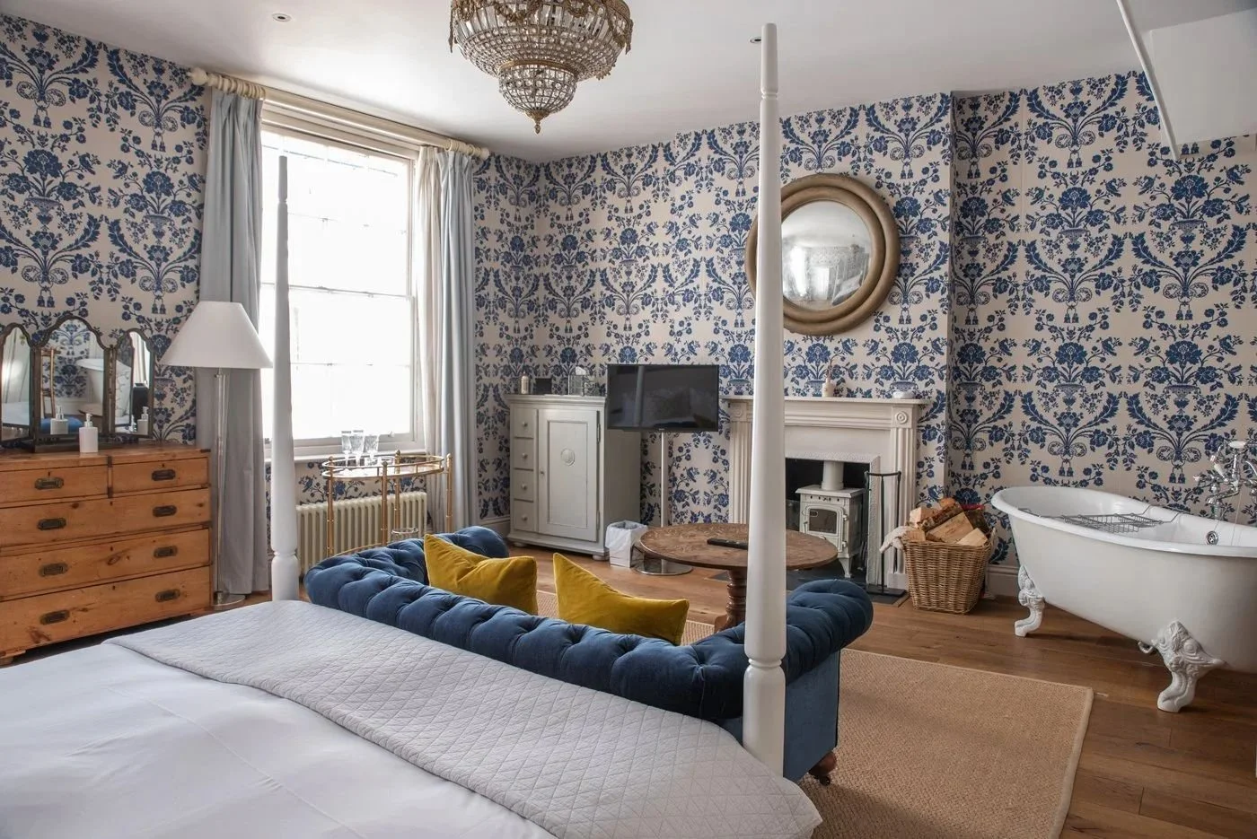 A cozy bedroom with blue and white floral wallpaper, a blue tufted sofa with yellow pillows, and a white clawfoot bathtub in the corner.
