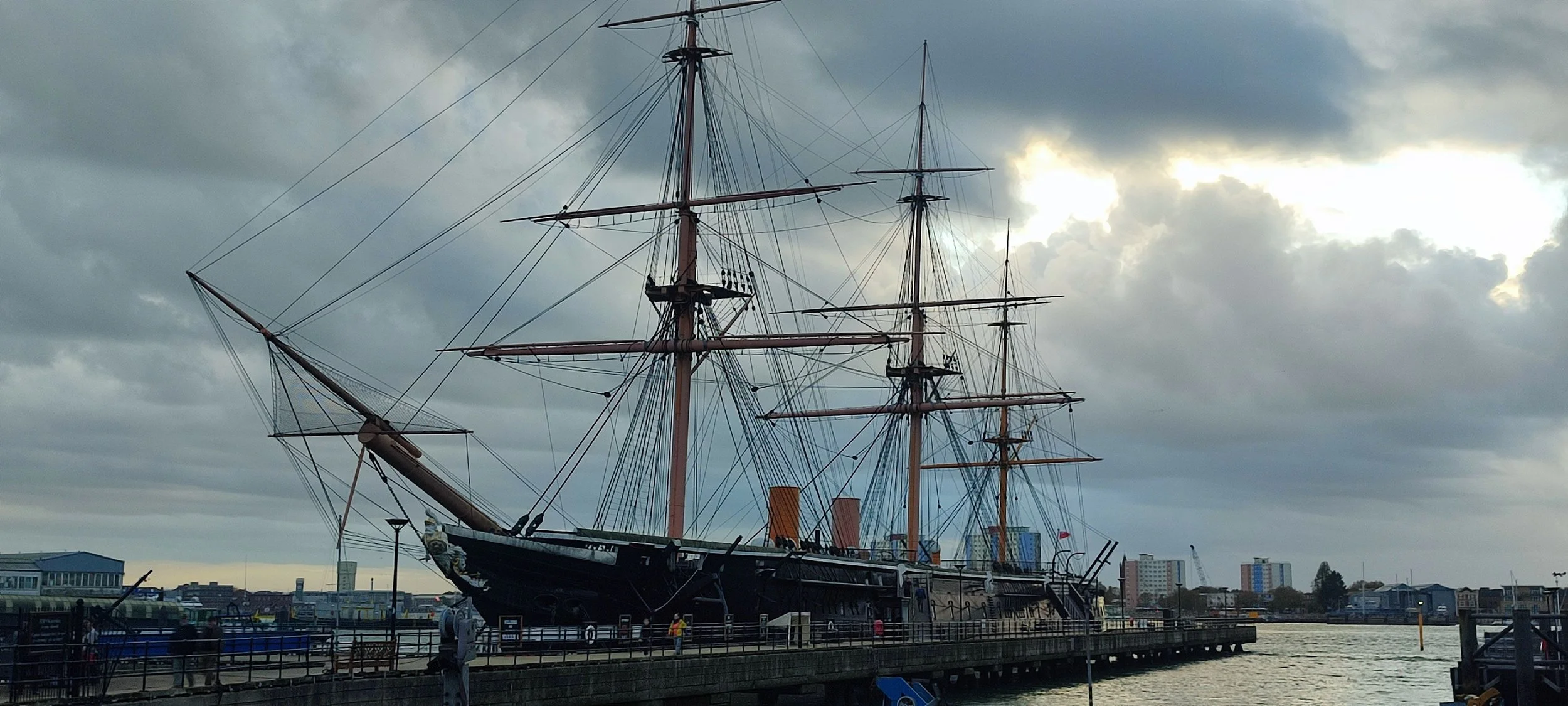 Historic tall ship with three masts docked at a waterfront pier under cloudy sky.