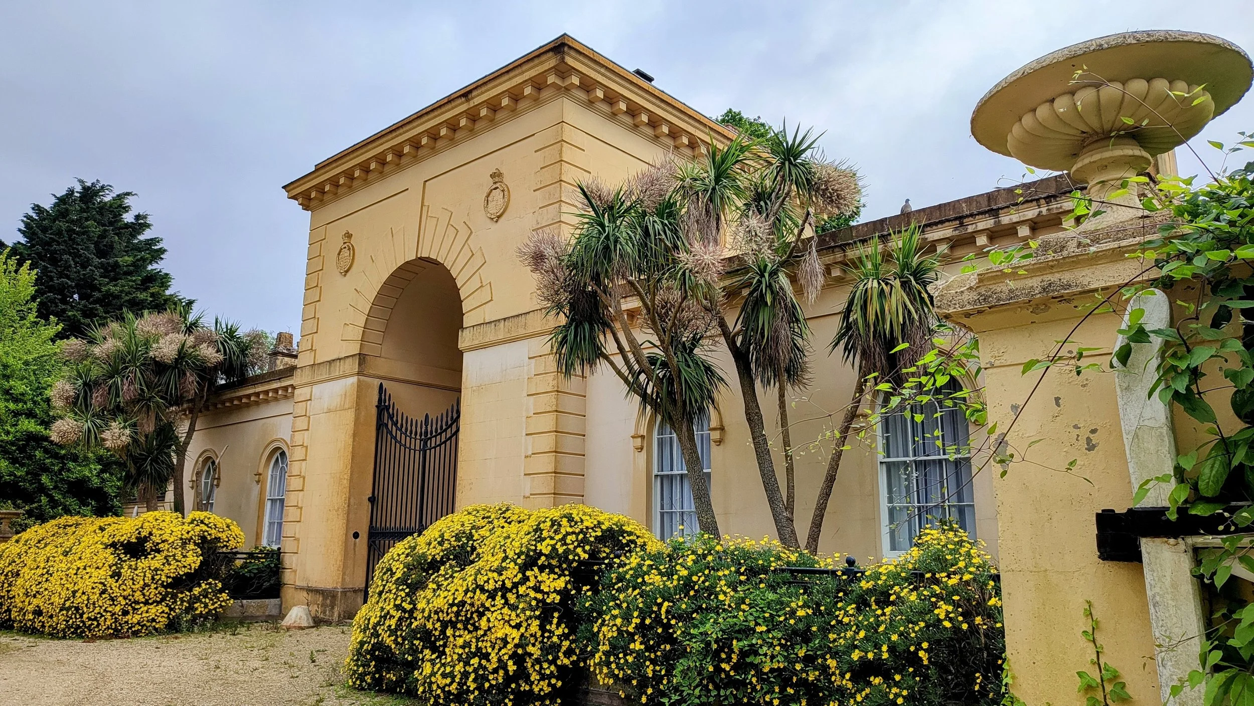 Yellow Victorian-style house with arched doorways, black iron gate, and lush garden with yellow flowers, green bushes, and trees.