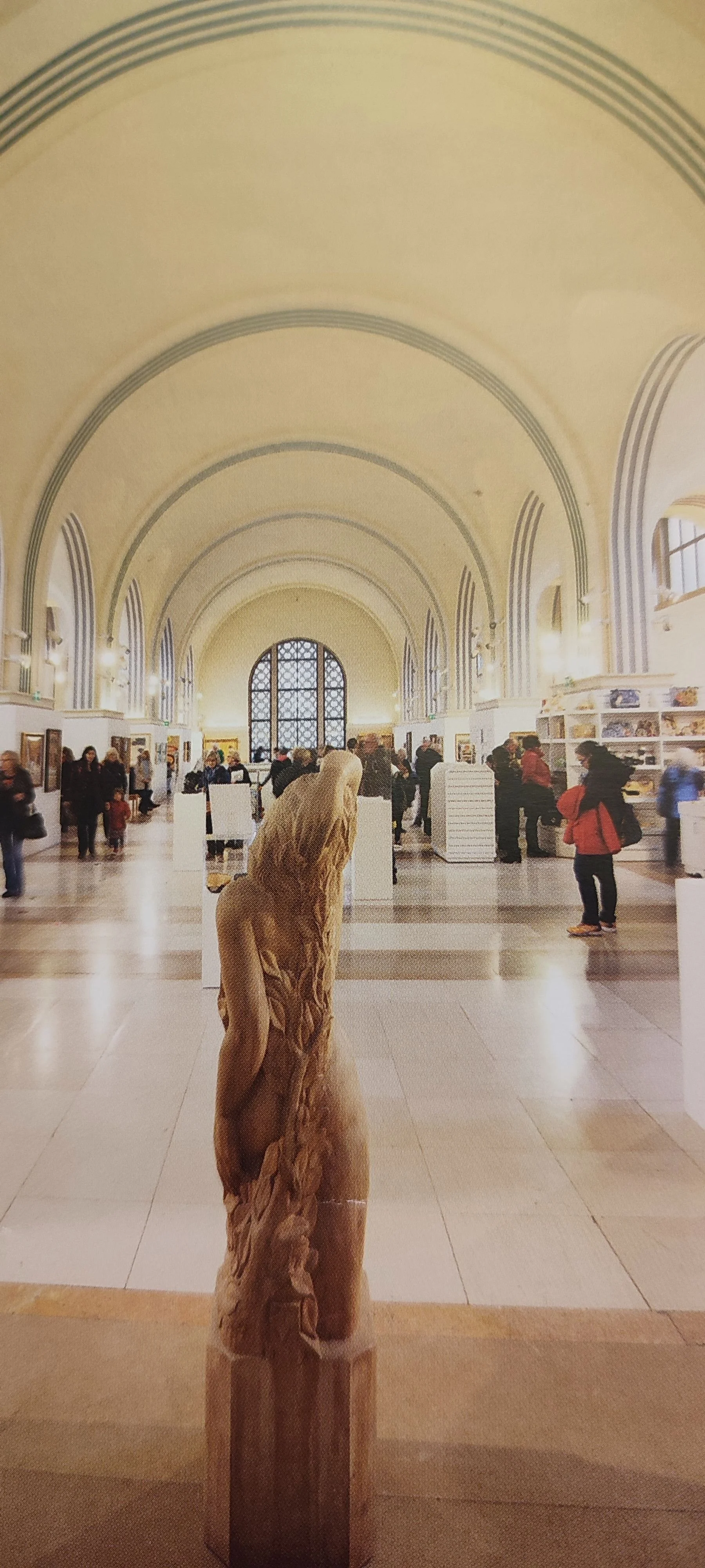 A museum interior with high arched ceilings, large window, and visitors viewing artwork; a wooden sculpture of a woman standing on a pedestal in the foreground.