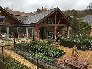 A rustic house with a garden in front, outdoor seating, and people inside visible through large windows.
