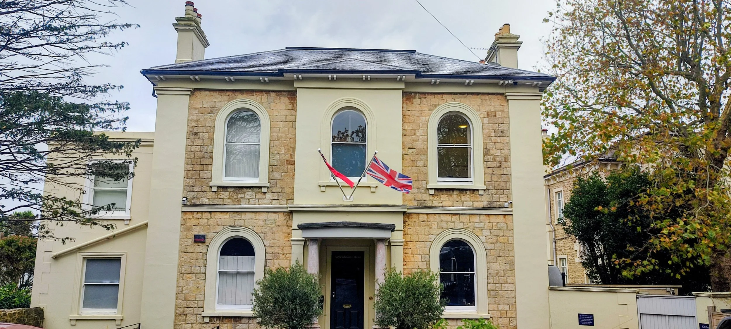 A two-story house with stone and cream-colored exterior walls, arched windows, and two flags (a Union Jack and UK Red Ensign) hanging from the second-floor balcony. The house is surrounded by trees with autumn leaves.