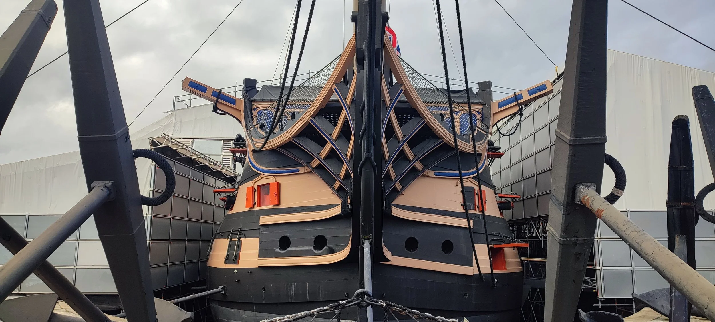 View of a ship's hull under construction with scaffolding and support beams around it, showing the ship's bow and detailed wooden framework.