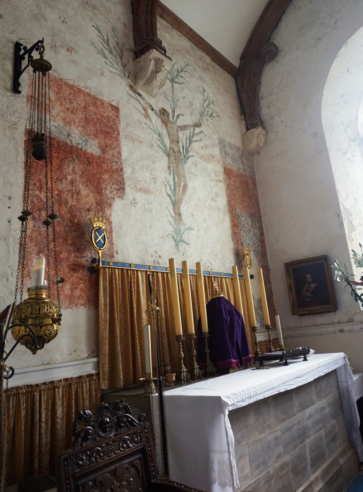 Interior of a church or chapel with an altar, gold and purple drapes, candles, and religious paintings on the wall. Faded fresco of a tree on the wall above the altar.